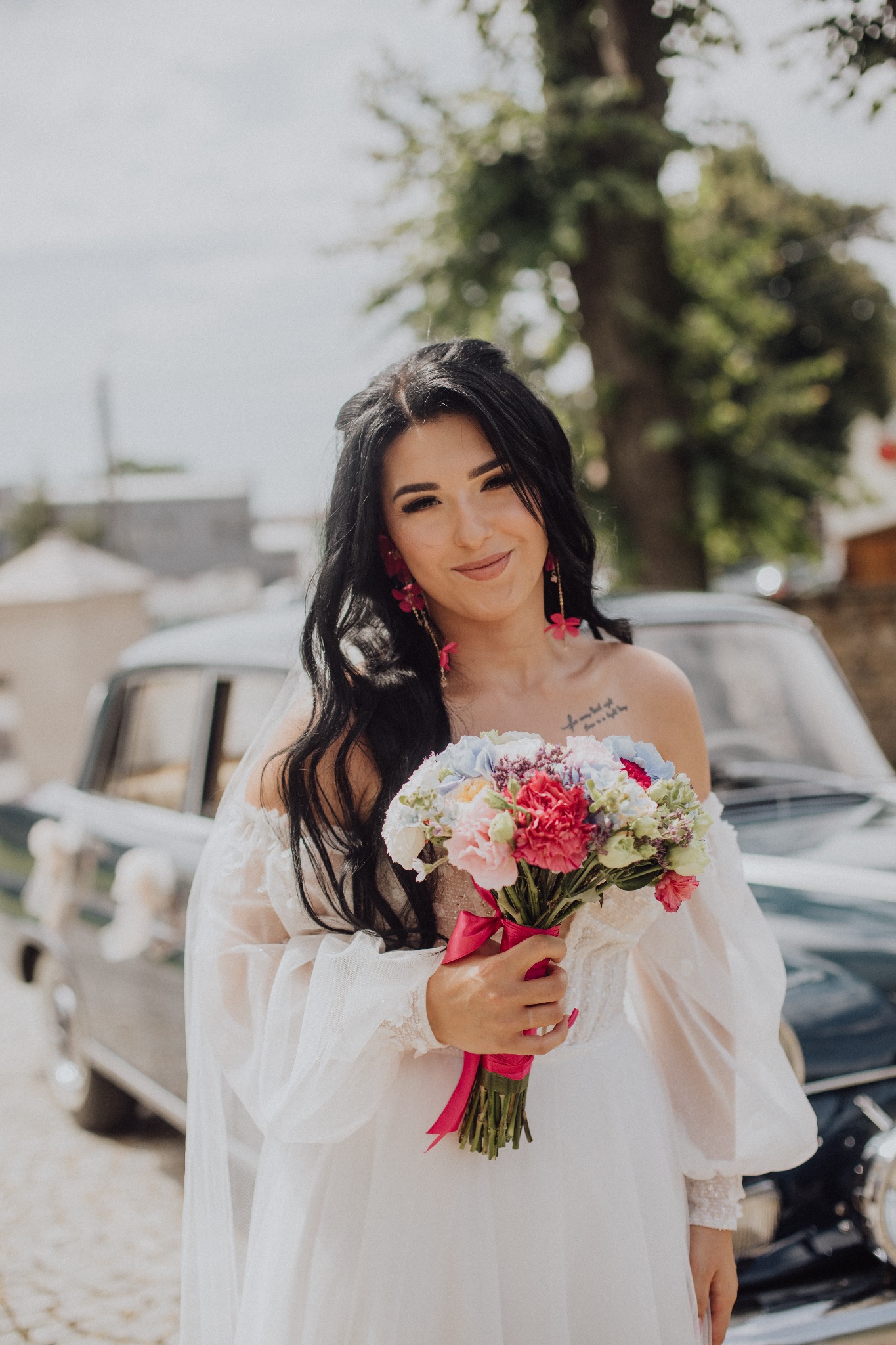 Uśmiechnięta panna młoda w białej sukni, trzymająca bukiet kwiatów z czerwoną wstążką, na tle klasycznego samochodu i drzewa.