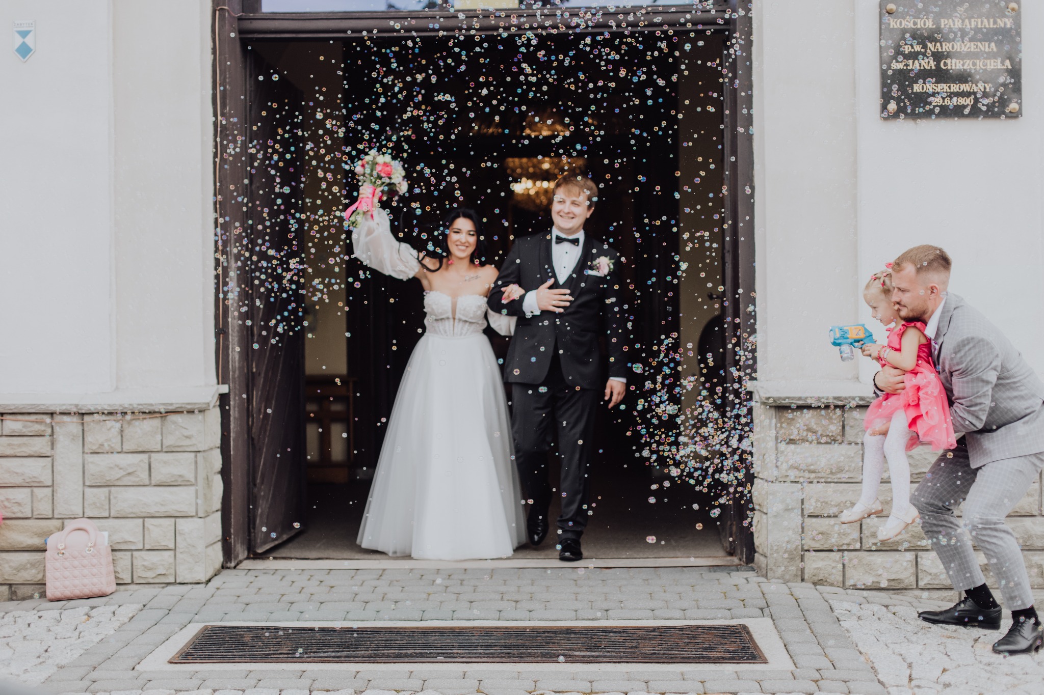 Uśmiechnięta para młoda wychodzi z kościoła, otoczona bańkami mydlanymi. W tle budynek kościoła i mężczyzna z dzieckiem puszczający bańki.