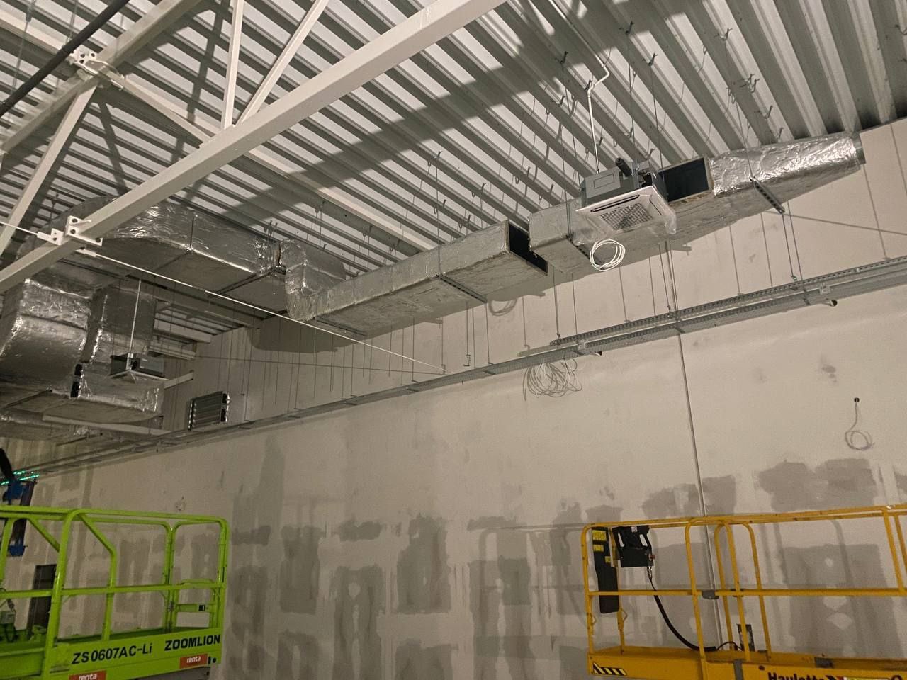 Instalacja kanałów wentylacyjnych z izolacją termiczną, widoczne podnośniki nożycowe, surowy stan ścian i konstrukcja stalowa dachu.