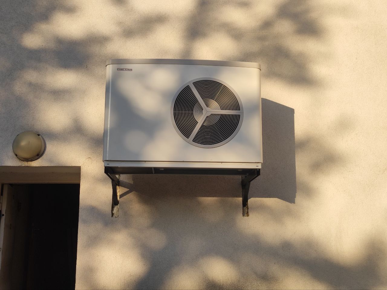 Zewnętrzna jednostka klimatyzacji marki Stiebel Eltron zamontowana na wspornikach na ścianie budynku, oświetlona popołudniowym słońcem rzucającym cienie liści.