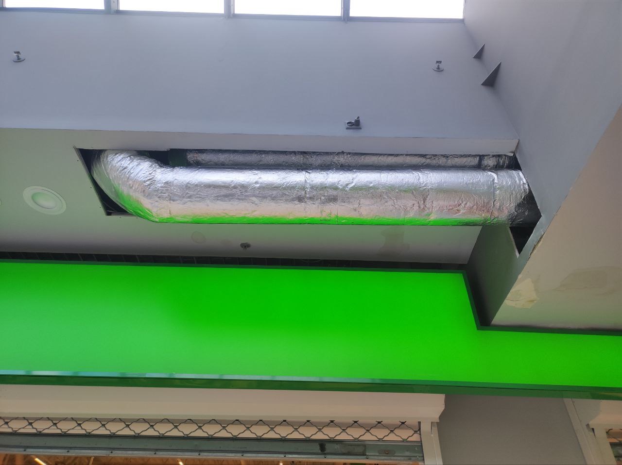Izolowany kanał wentylacyjny, pokryty folią aluminiową, zamontowany pod sufitem w przestrzeni komercyjnej, widoczny nad jaskrawozieloną powierzchnią.