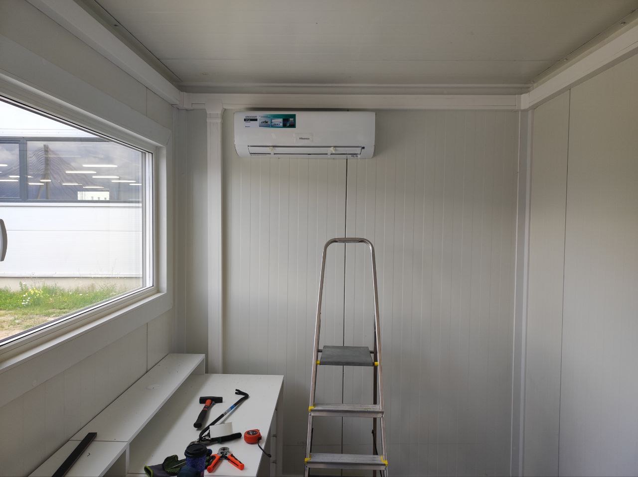 Wnętrze pomieszczenia z zamontowaną klimatyzacją Hisense, widoczna aluminiowa drabina, stół z narzędziami, okno z widokiem na budynek przemysłowy.