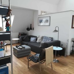 Usługi Remontowo Budowlane - Jasne wnętrze pokoju z drewnianą podłogą, szarą sofą, czarnym krzesłem z beżowym pledem, regałem z książkami i drabinką opartą o ścianę, poziomica oparta o ścianę obok świeczników.