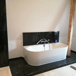 Usługi Remontowo Budowlane - Nowoczesna łazienka z wolnostojącą białą wanną na tle czarnej marmurowej ściany i podłogi, drewniany słup konstrukcyjny w rogu pomieszczenia.