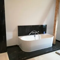 Nowoczesna łazienka z wolnostojącą białą wanną na tle czarnej marmurowej ściany i podłogi, drewniany słup konstrukcyjny w rogu pomieszczenia.