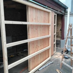 Usługi Remontowo Budowlane - Drewniane przesuwne drzwi w trakcie montażu, widoczne prowadnice, rama z listew, surowy stan drewna, drabina oparta o ścianę, narzędzia na podłodze.