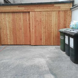 Usługi Remontowo Budowlane - Nowa, drewniana brama garażowa przesuwna, wykonana z pionowo ułożonych desek, z widocznym fragmentem pomarańczowego kabla na szarym podłożu i pojemnikami na śmieci obok.