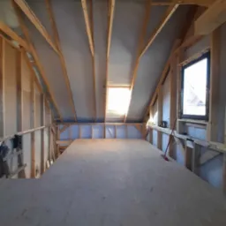 Poddasze w trakcie adaptacji: widoczna konstrukcja drewniana, izolacja, okno dachowe i okno w ścianie szczytowej, przygotowana podłoga z płyt OSB, czerwona zmiotka oparta o ścianę.