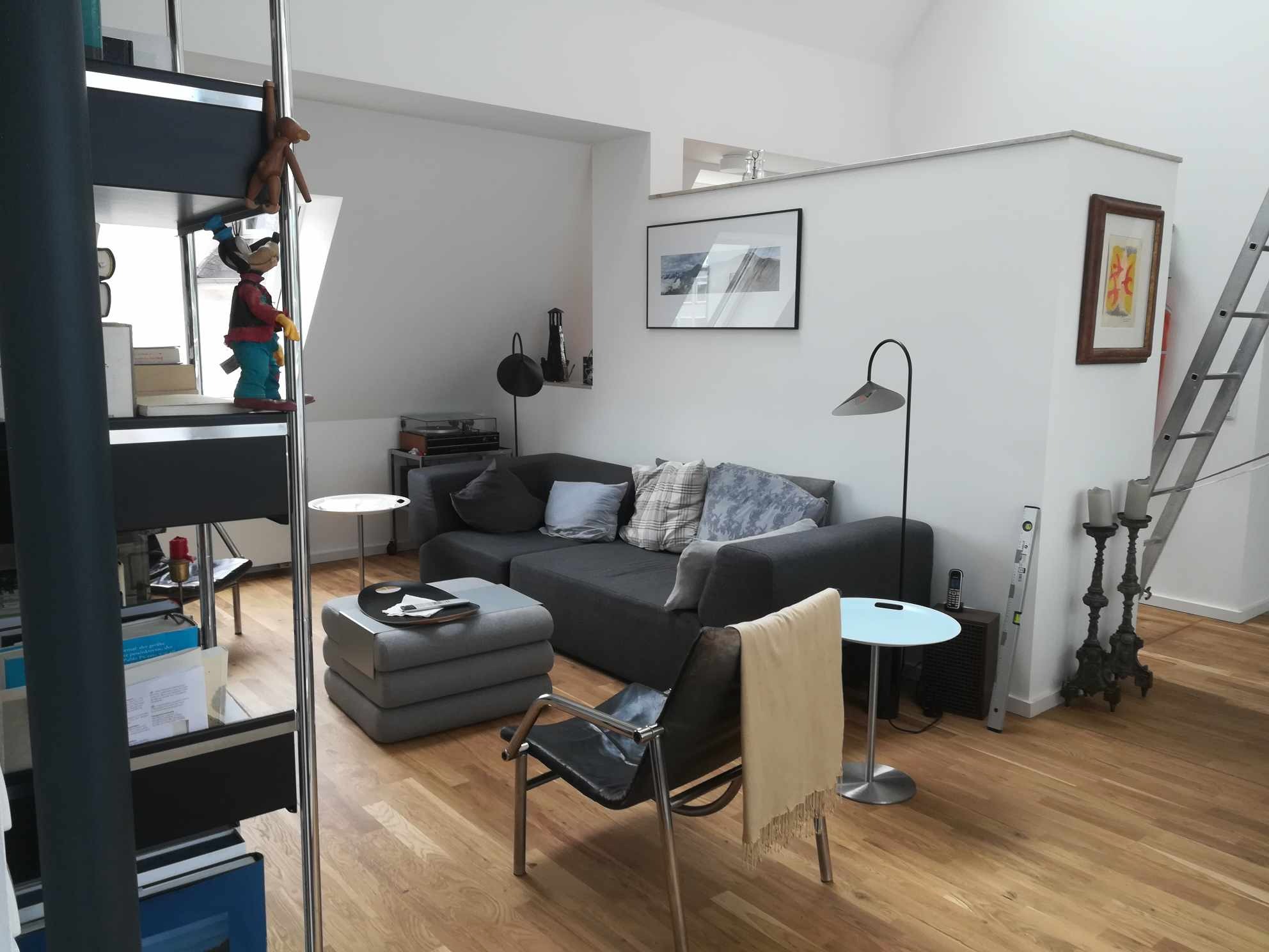 Jasne wnętrze pokoju z drewnianą podłogą, szarą sofą, czarnym krzesłem z beżowym pledem, regałem z książkami i drabinką opartą o ścianę, poziomica oparta o ścianę obok świeczników.