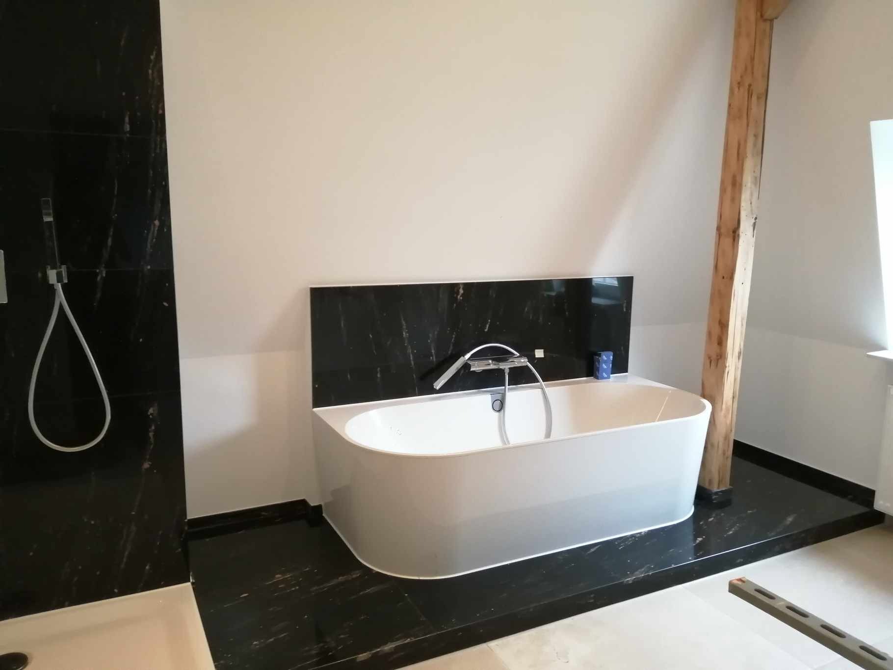 Nowoczesna łazienka z wolnostojącą białą wanną na tle czarnej marmurowej ściany i podłogi, drewniany słup konstrukcyjny w rogu pomieszczenia.