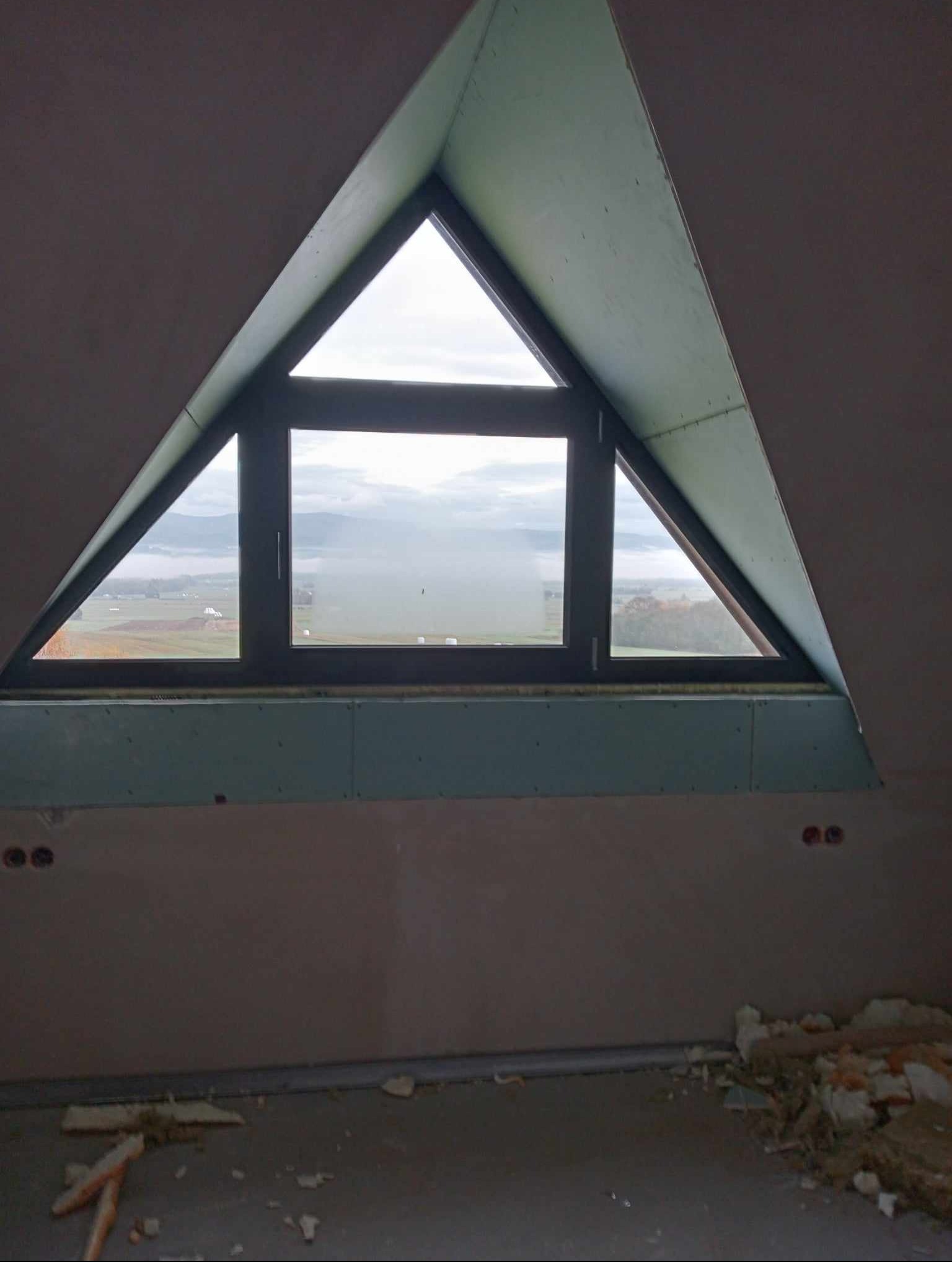 Poddasze w trakcie adaptacji: trójkątne okno dachowe z widokiem na krajobraz, ściany wykończone płytami gipsowo-kartonowymi, widoczne puszki elektryczne i resztki materiałów budowlanych na podłodze.