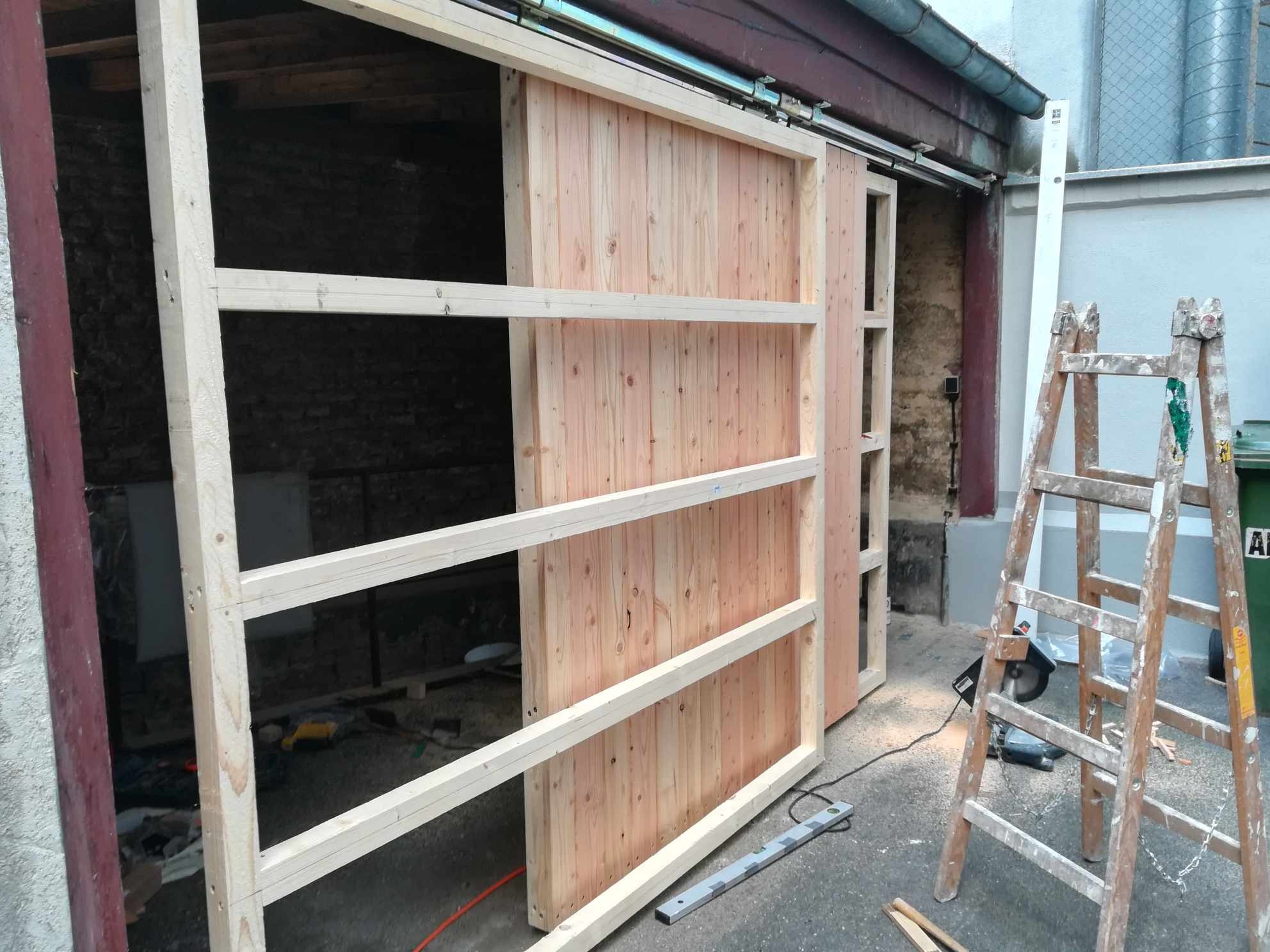 Drewniane przesuwne drzwi w trakcie montażu, widoczne prowadnice, rama z listew, surowy stan drewna, drabina oparta o ścianę, narzędzia na podłodze.