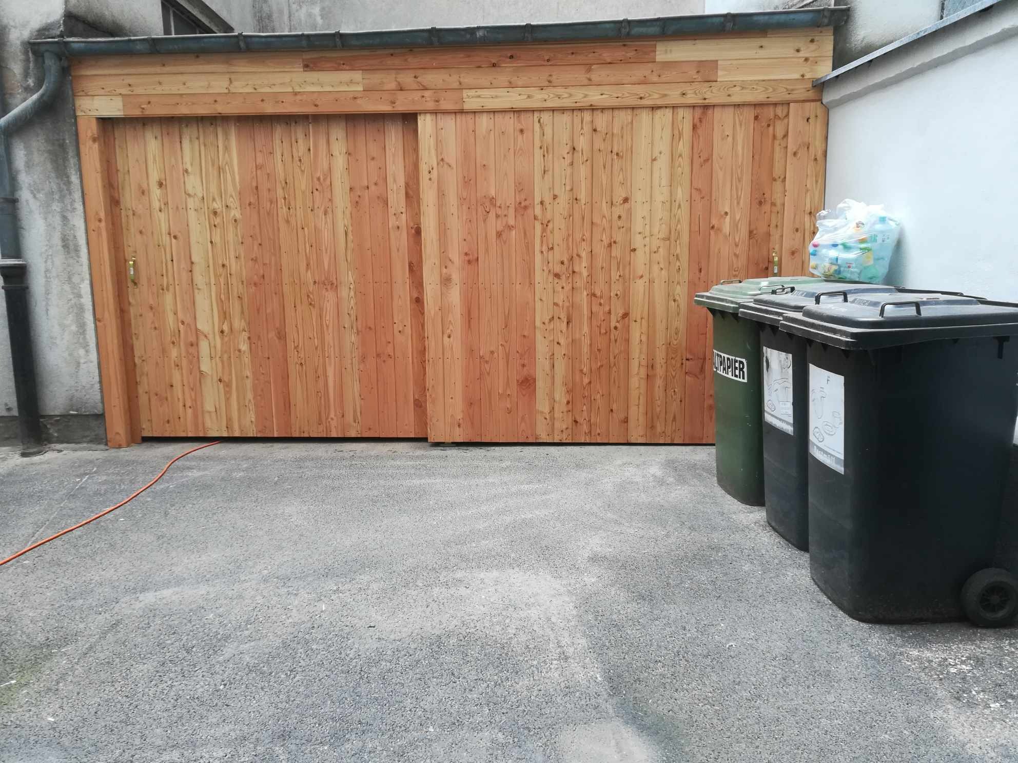 Nowa, drewniana brama garażowa przesuwna, wykonana z pionowo ułożonych desek, z widocznym fragmentem pomarańczowego kabla na szarym podłożu i pojemnikami na śmieci obok.