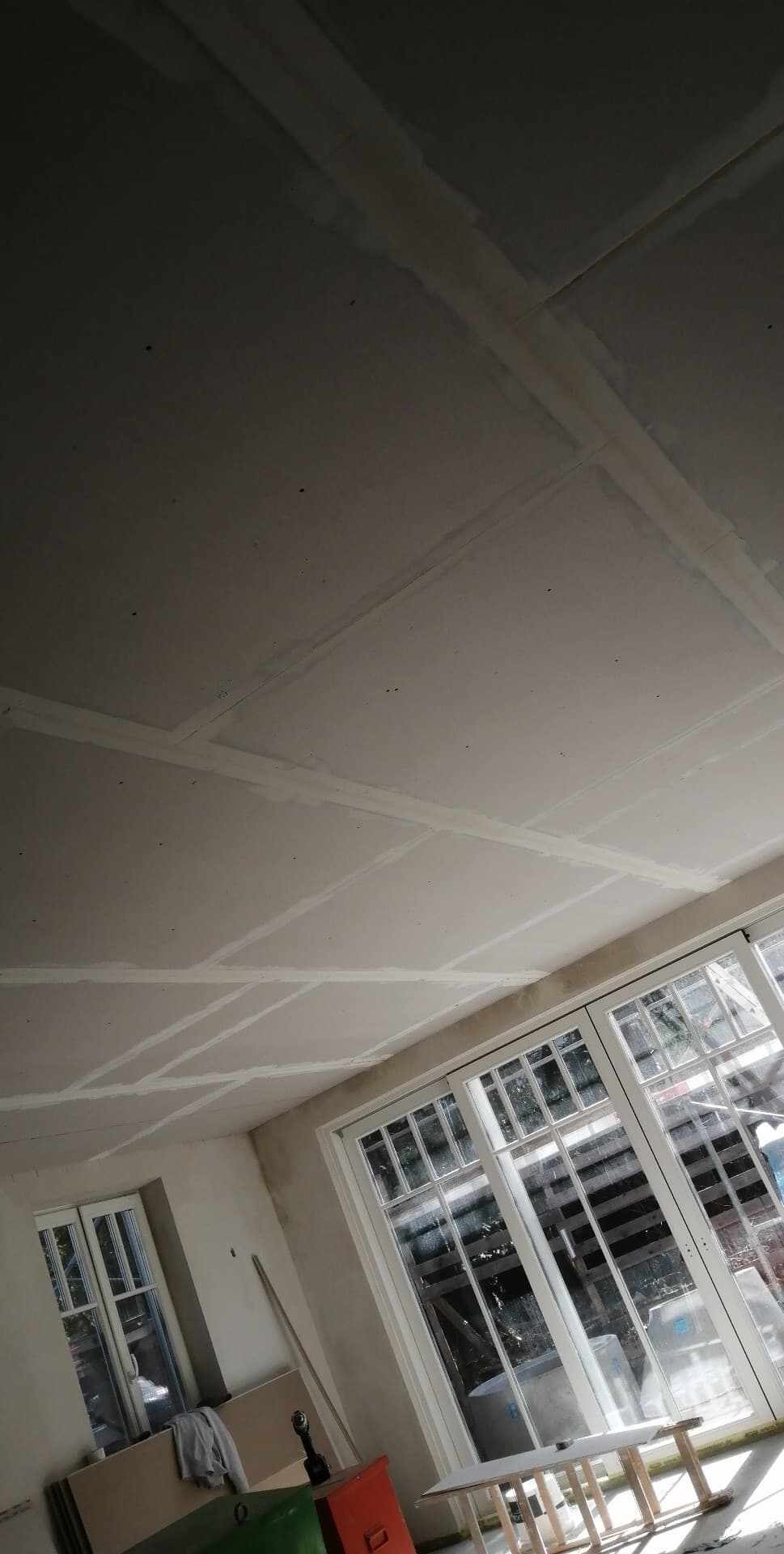 Sufit z płyt kartonowo-gipsowych w trakcie szpachlowania łączeń, widoczne okna balkonowe z białymi szprosami i fragmenty narzędzi na podłodze.
