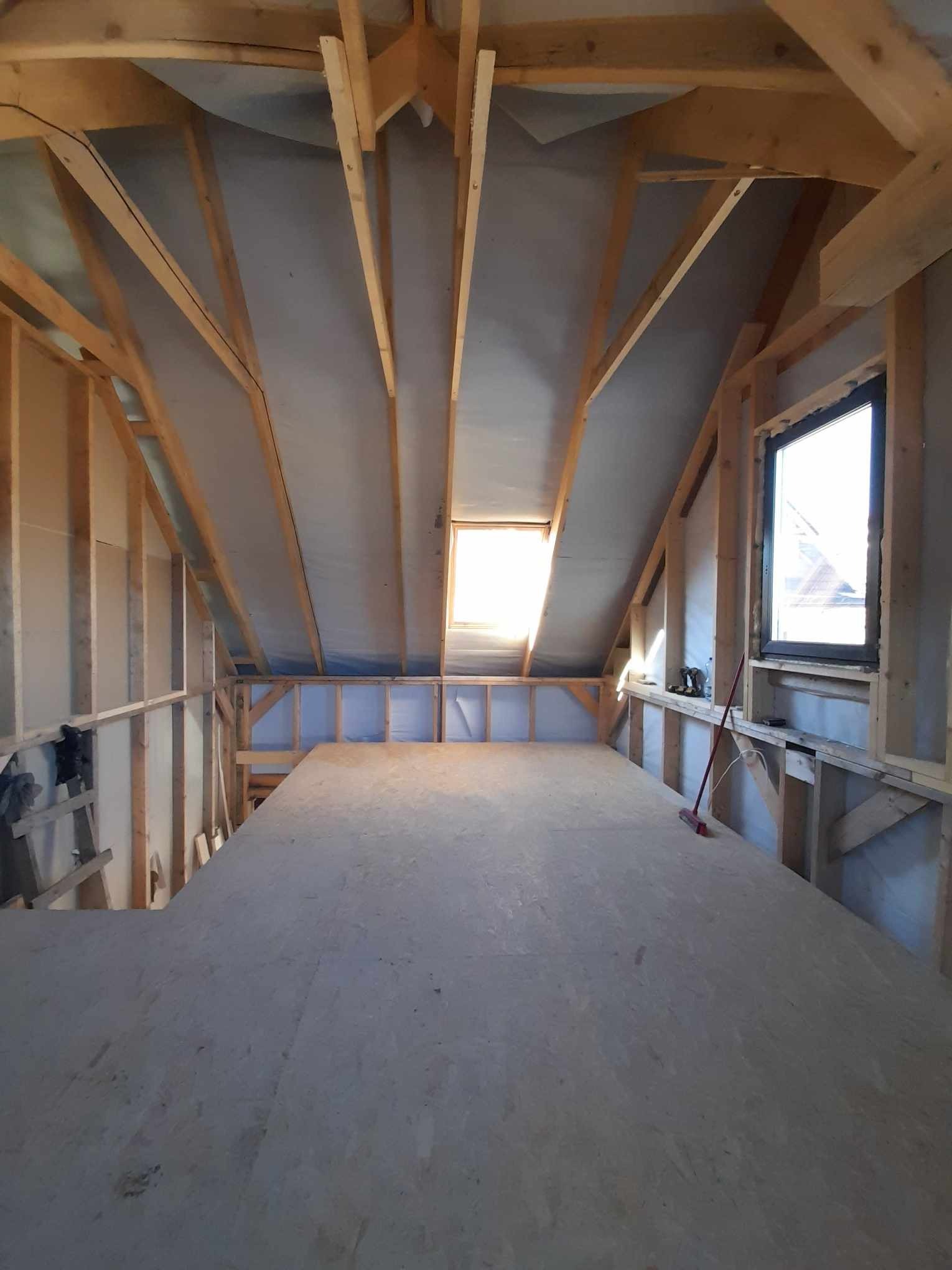 Poddasze w trakcie adaptacji: widoczna konstrukcja drewniana, izolacja, okno dachowe i okno w ścianie szczytowej, przygotowana podłoga z płyt OSB, czerwona zmiotka oparta o ścianę.