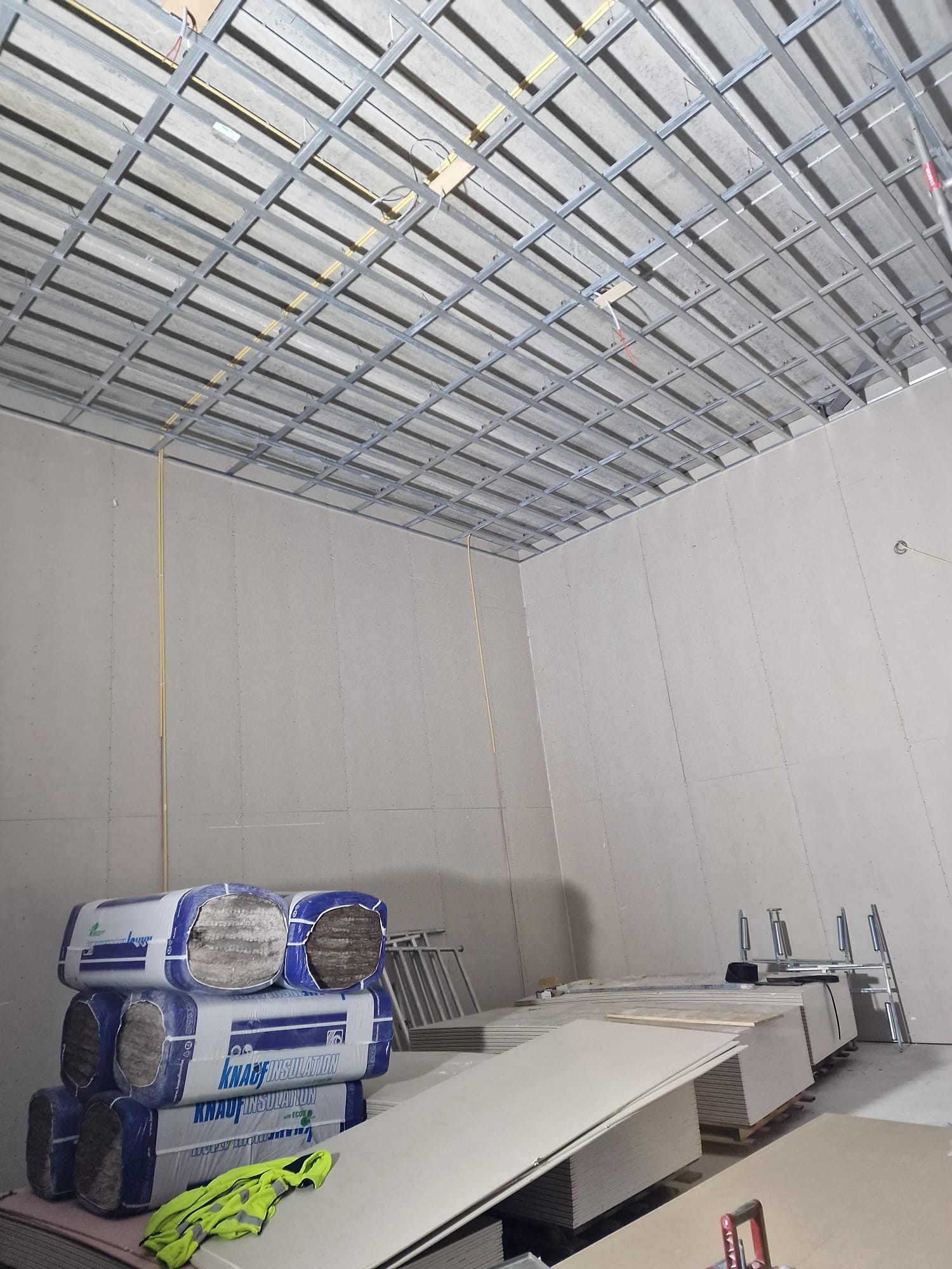 Wnętrze pomieszczenia w trakcie prac wykończeniowych: widoczna metalowa konstrukcja sufitu podwieszanego, ściany pokryte płytami oraz stosy materiałów izolacyjnych Knauf Insulation i płyt...