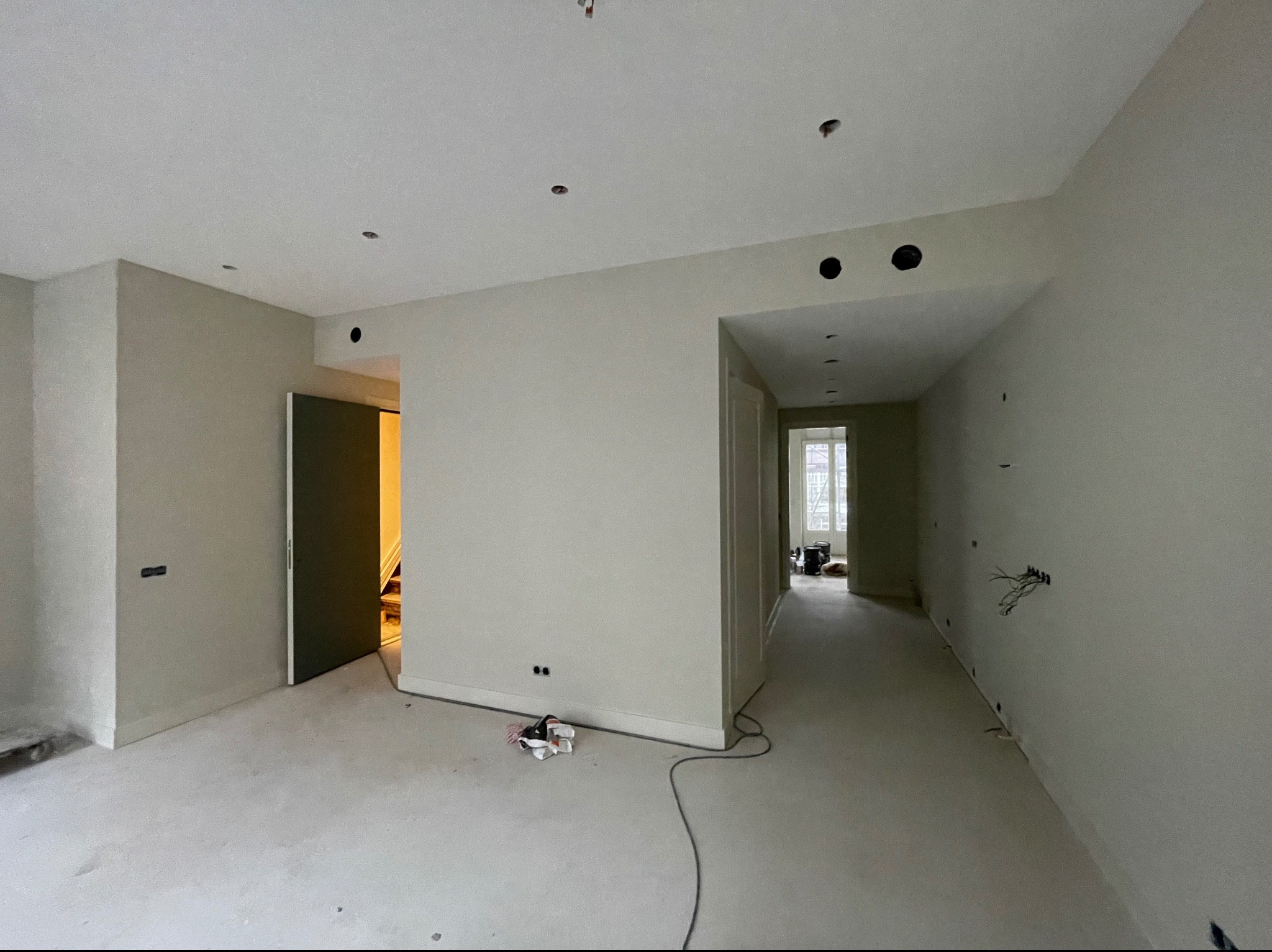 Wnętrze w trakcie remontu: surowe ściany w odcieniach beżu, przygotowane otwory pod oświetlenie w suficie, widoczne przewody elektryczne, podłoga w trakcie wylewki, częściowo zasłonięte drzwi...