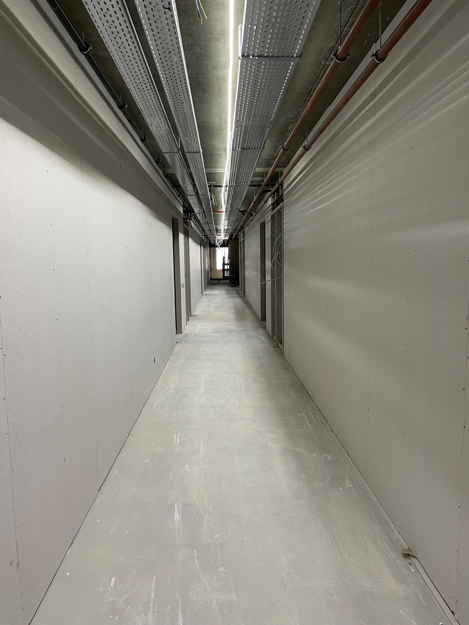 Długi korytarz w surowym stanie wykończenia, ściany pokryte szarą płytą, widoczne instalacje pod sufitem, perspektywa zbieżna w kierunku odległego okna.