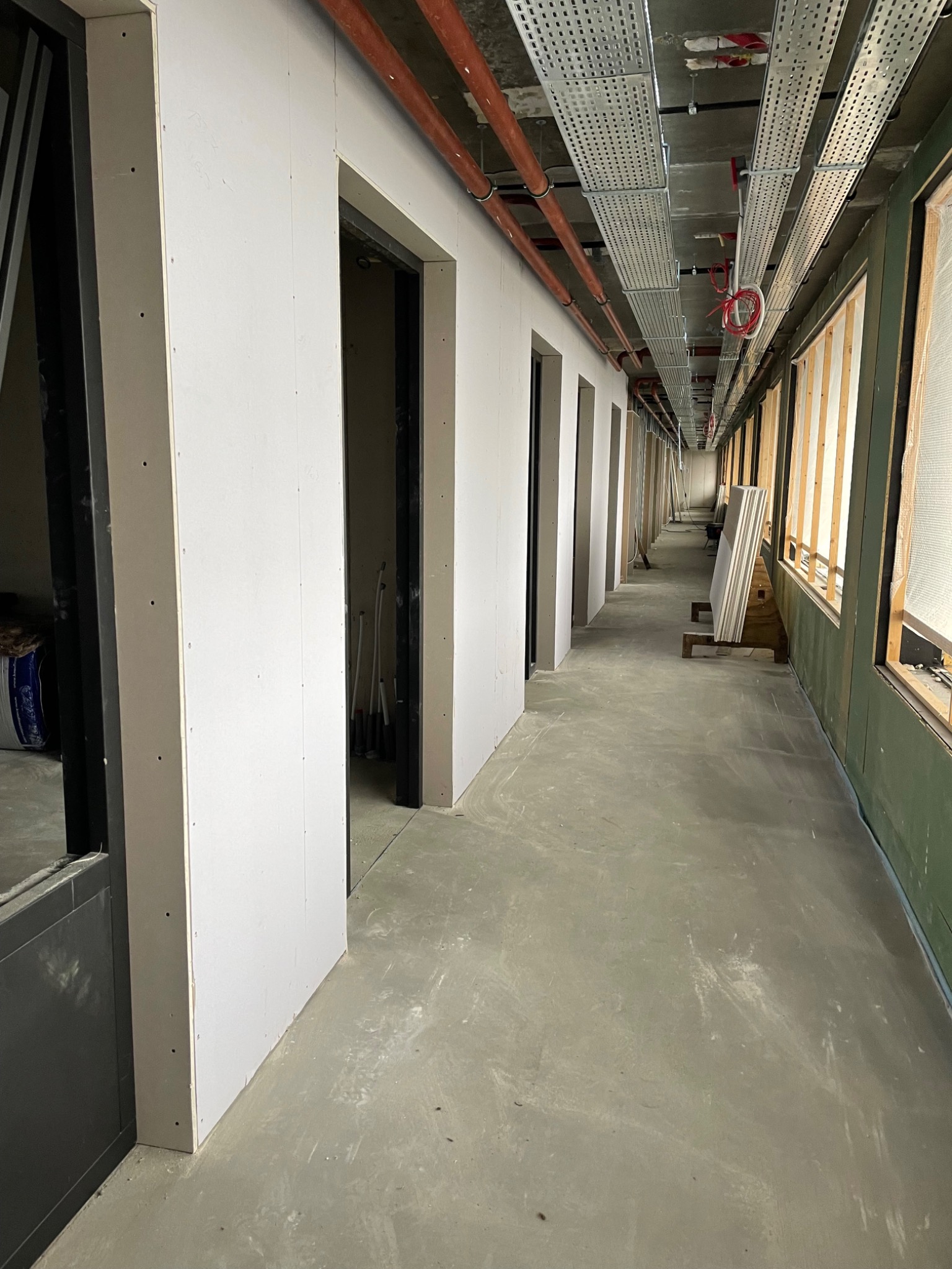 Korytarz w trakcie wykańczania: ściany z płyt z widocznymi otworami po wkrętach, metalowe korytka kablowe pod sufitem, surowe podłoże betonowe, okna w drewnianych ramach z zielonymi elementami.