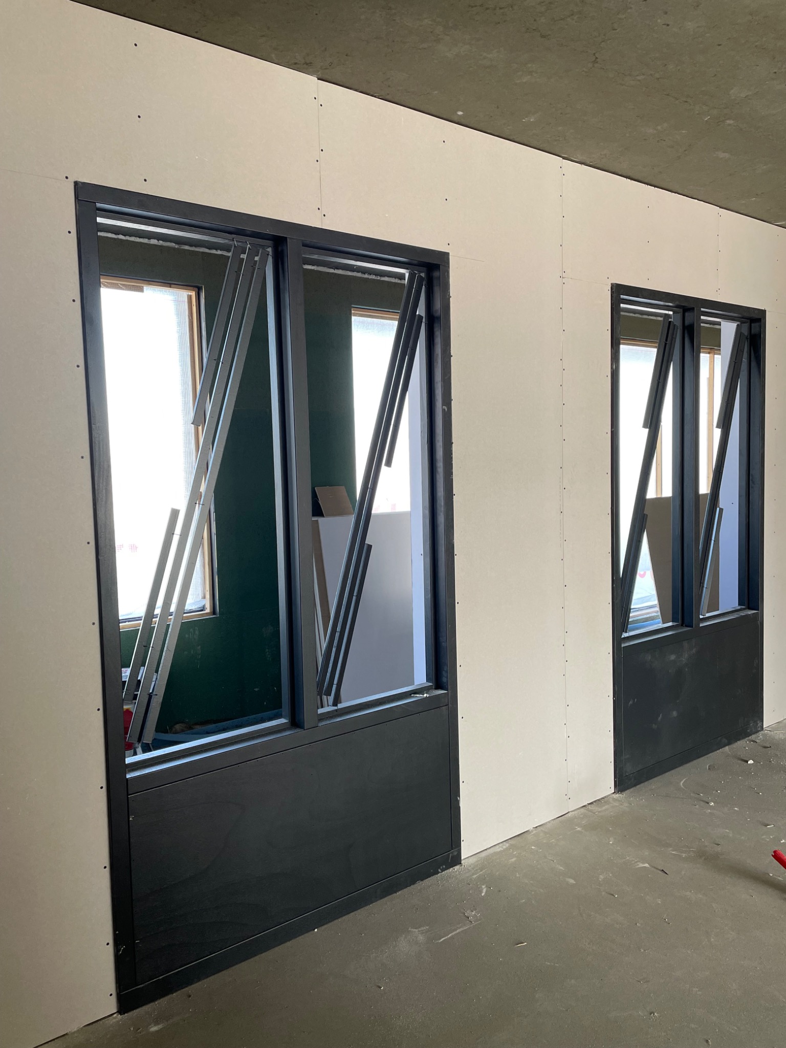 Montaż ścian działowych: widoczne dwie czarne ramy okienne z częściowo zamontowanymi, ukośnymi żaluzjami w ścianie z płyt gipsowo-kartonowych, surowy betonowy sufit i szara podłoga w trakcie prac...