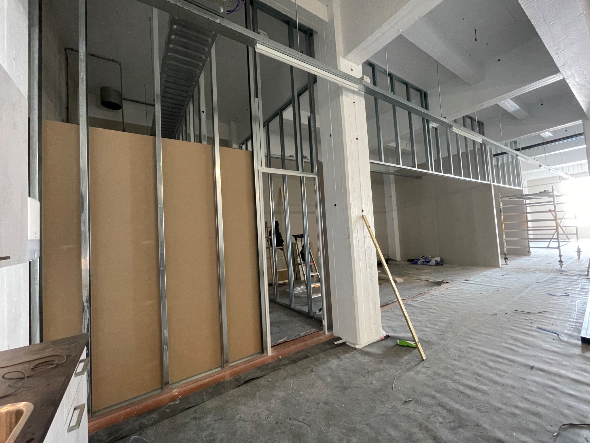 Montaż metalowej konstrukcji ścian działowych z brązowymi panelami w surowym wnętrzu budynku, widoczne kable i elementy instalacji pod sufitem.