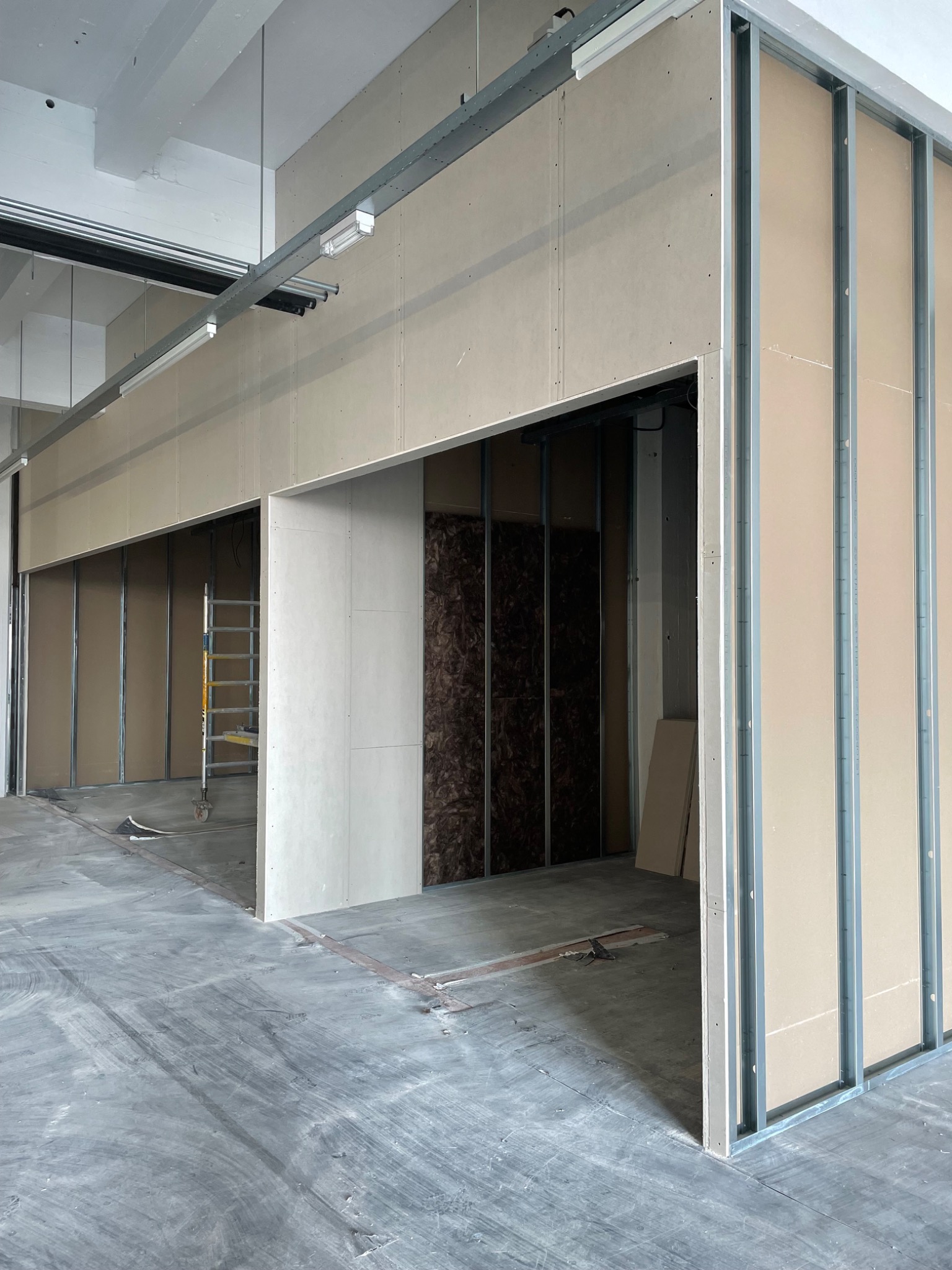 Montaż ścian działowych z płyty karton gips na metalowym stelażu, widoczna izolacja wewnątrz konstrukcji, surowy beton na podłodze i drabina w tle.