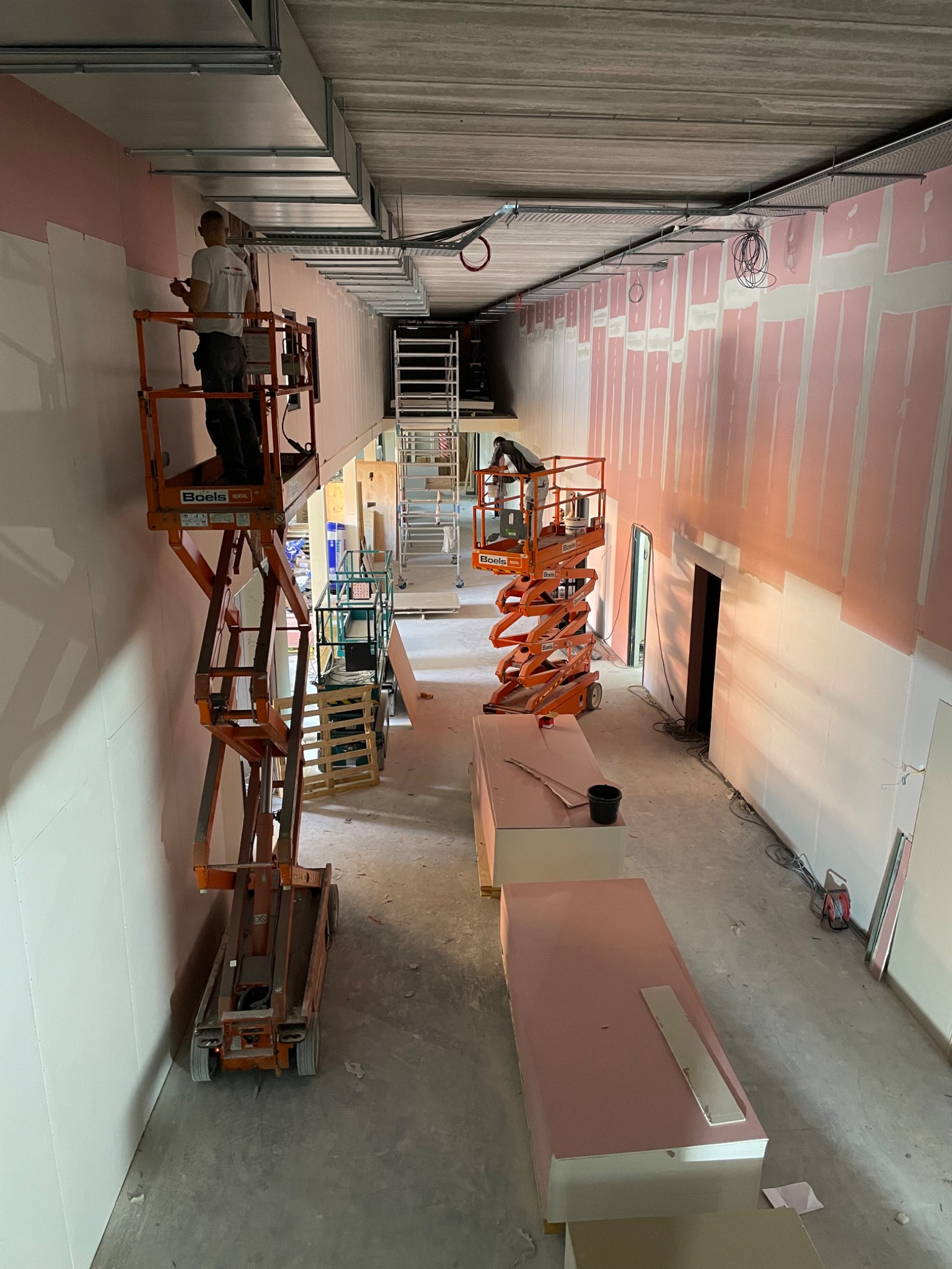 Montaż ścian działowych w długim pomieszczeniu z wykorzystaniem podnośników nożycowych, widoczne płyty z różowym podkładem i metalowe profile sufitowe.