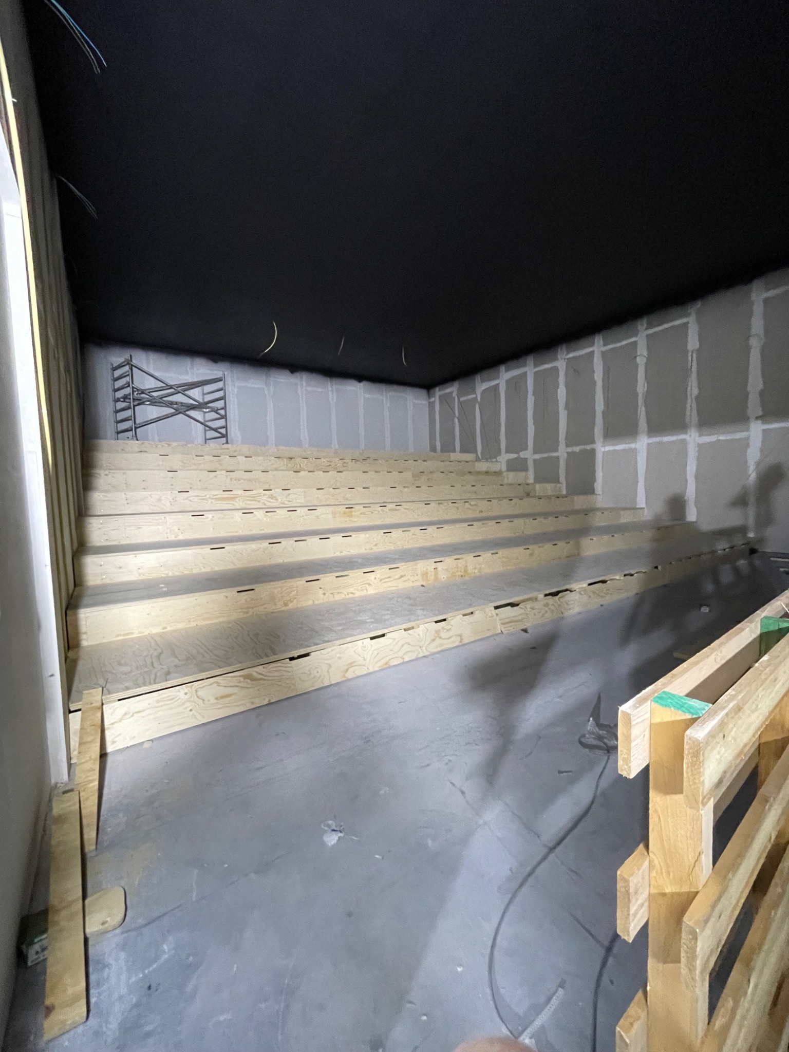 Konstrukcja schodkowa z drewna przygotowana w pomieszczeniu z surowymi ścianami i czarnym sufitem, widoczne kable i rusztowanie.