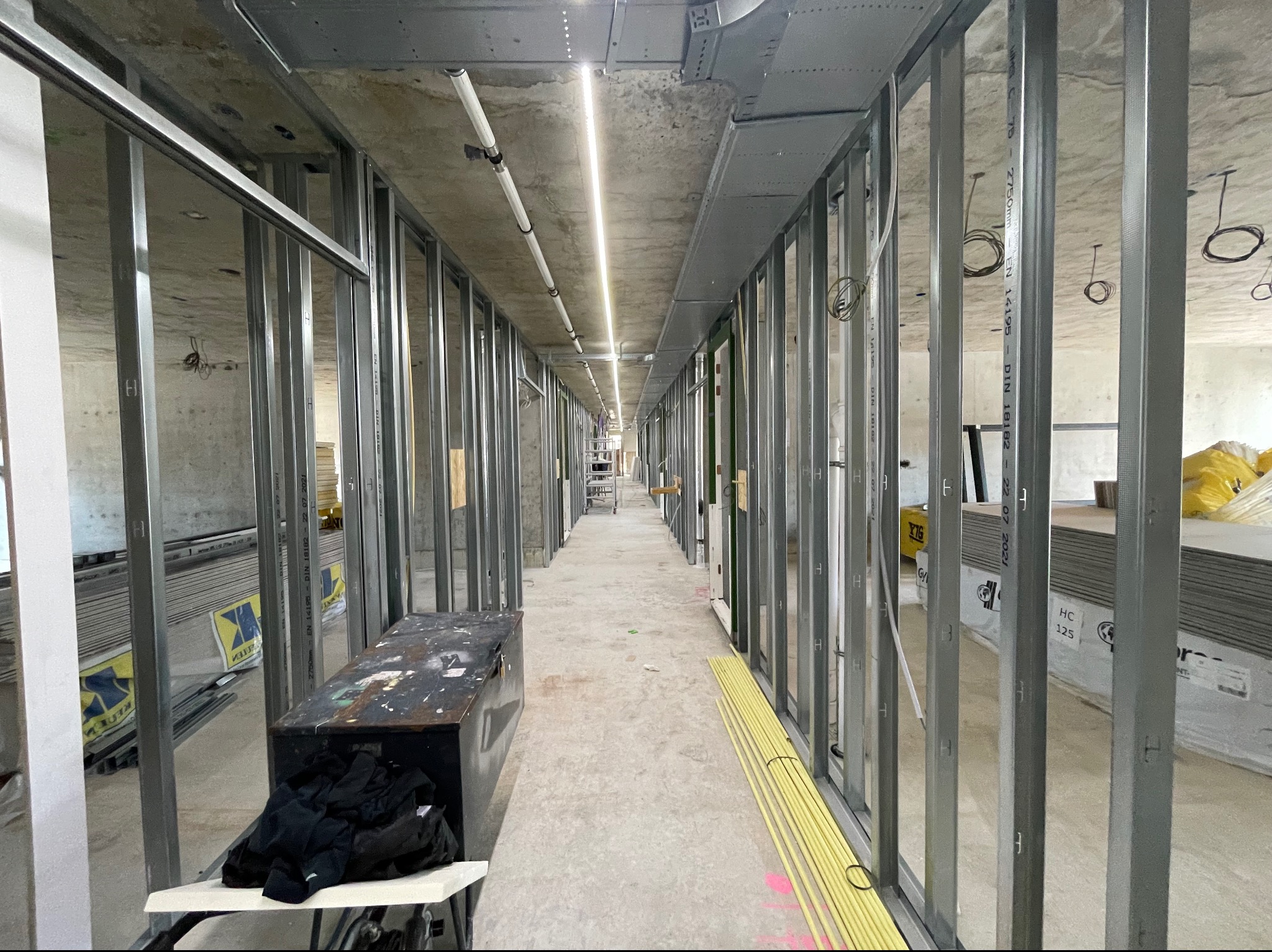 Długi korytarz w trakcie remontu, widoczne stalowe profile konstrukcyjne ścian działowych, pod sufitem instalacje, na podłodze materiały budowlane i skrzynia z narzędziami.