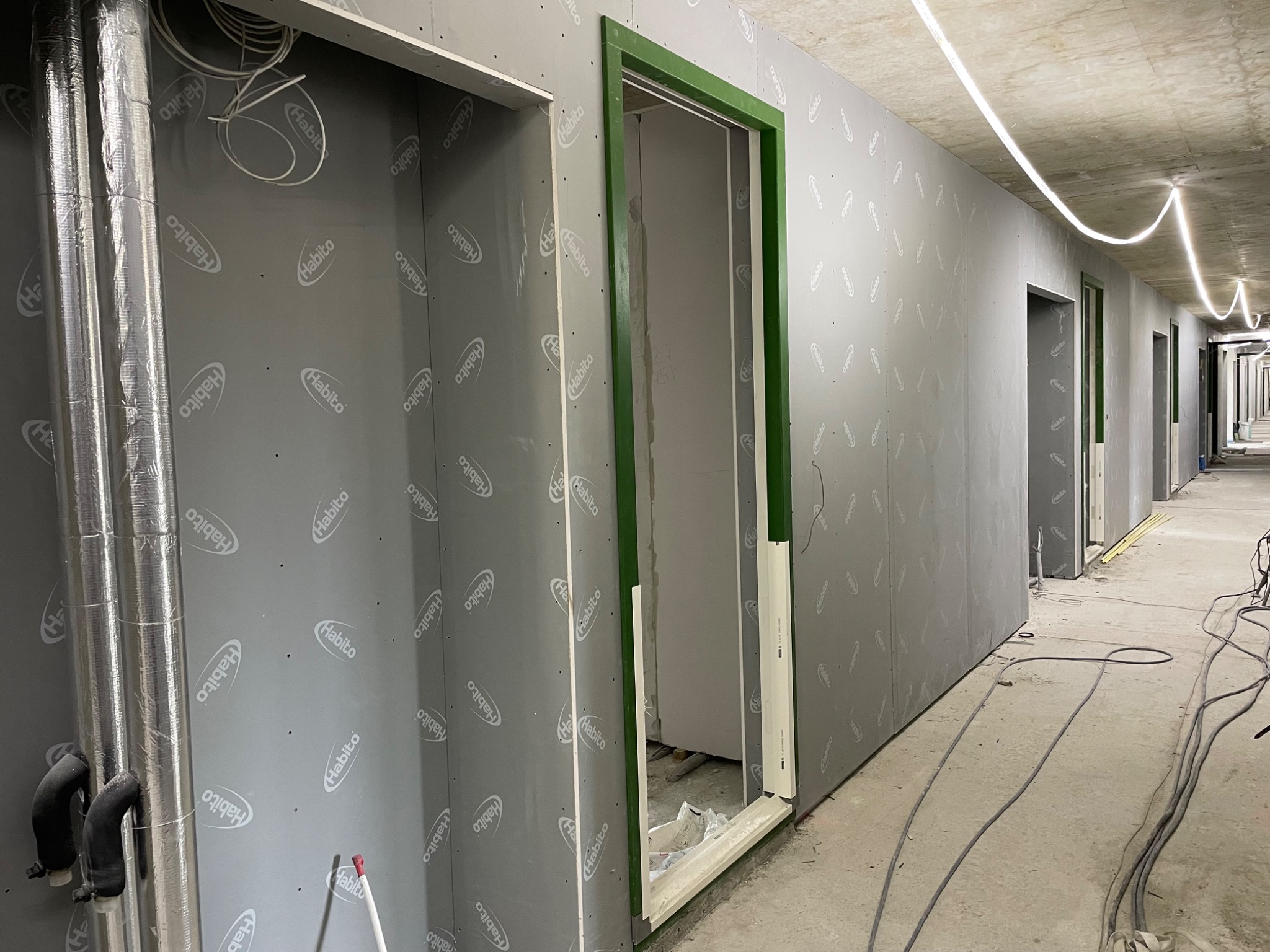 Korytarz w trakcie wykańczania, ściany obłożone szarymi płytami z widocznym logo producenta, zielone ramy drzwiowe, instalacje wentylacyjne i kable na podłodze.