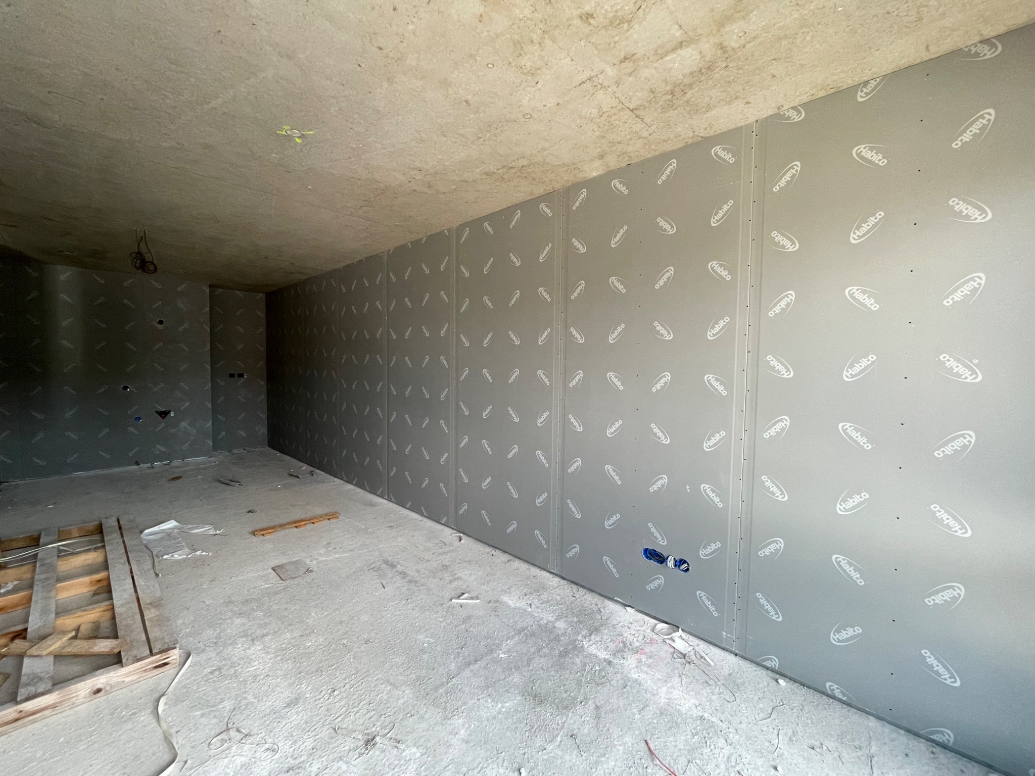 Montaż szarej płyty kartonowo-gipsowej Habito na ścianie w surowym, remontowanym pomieszczeniu z widoczną instalacją elektryczną i elementami drewnianej konstrukcji na podłodze.