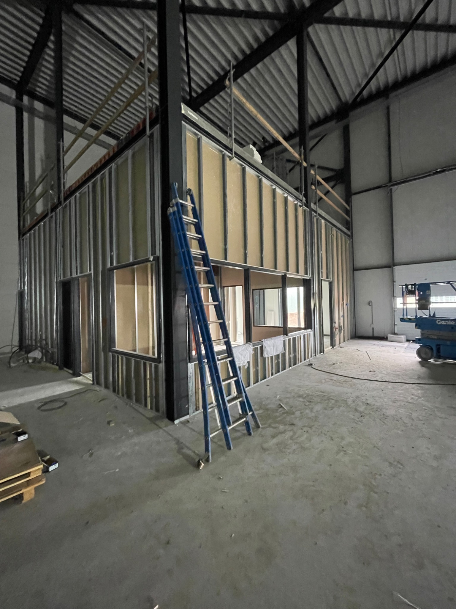 Wnętrze hali przemysłowej w trakcie tworzenia ścian działowych z metalowych profili i płyt wykończeniowych; widoczna niebieska drabina oparta o ścianę, podnośnik koszowy w tle.