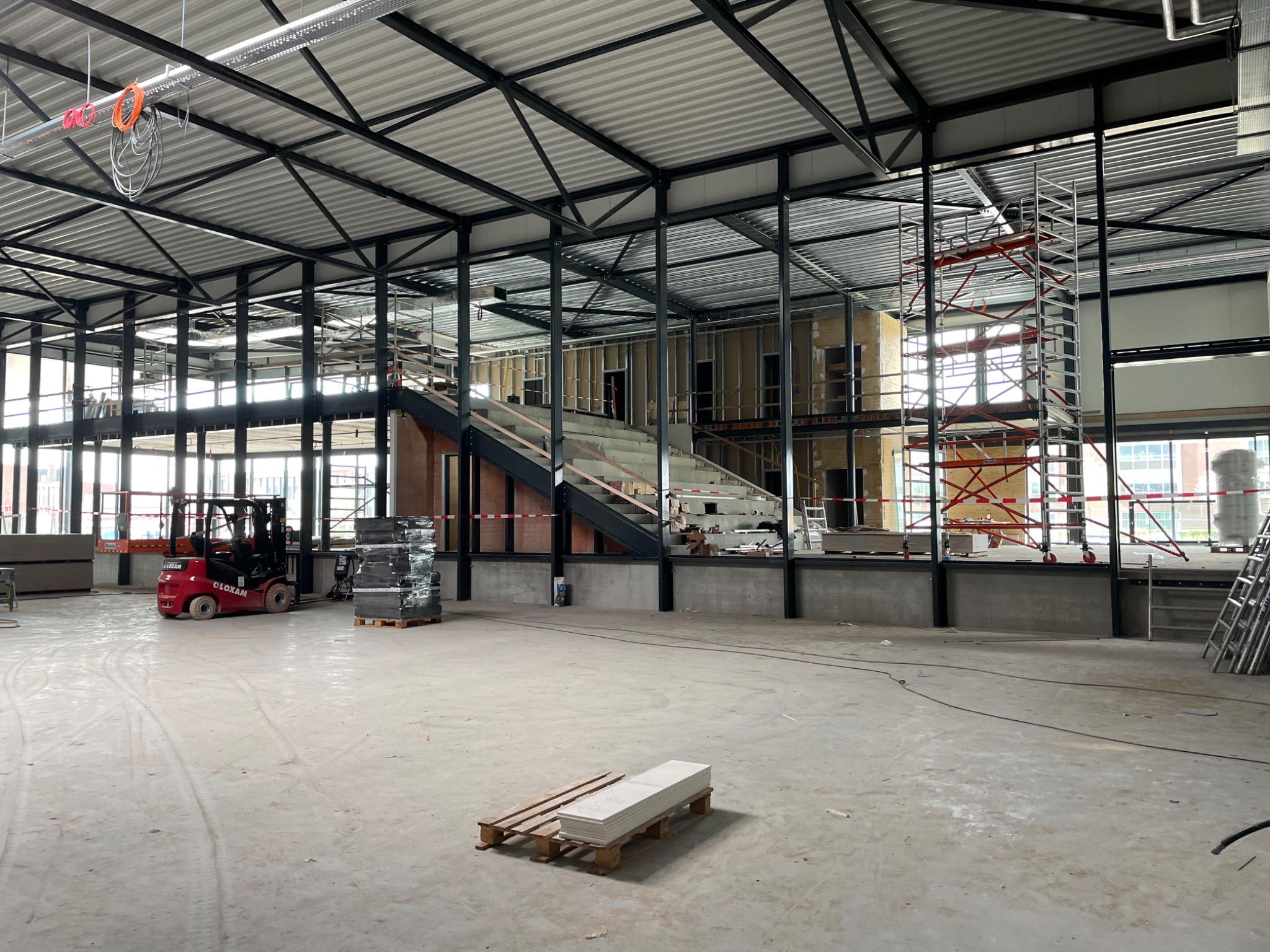 Wnętrze hali w trakcie prac wykończeniowych, widoczny szkielet konstrukcyjny, schody, podnośnik widłowy, palety z materiałami budowlanymi i rusztowania.
