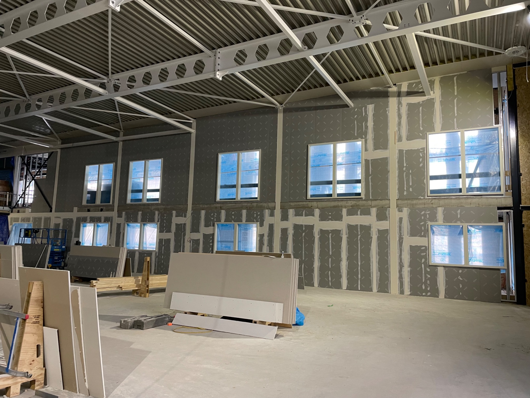 Wnętrze hali w trakcie prac wykończeniowych, ściany z zamontowanymi oknami pokryte szarą płytą, widoczne łączenia szpachlowane, na podłodze stos płyt i materiałów budowlanych, sufit z metalową...