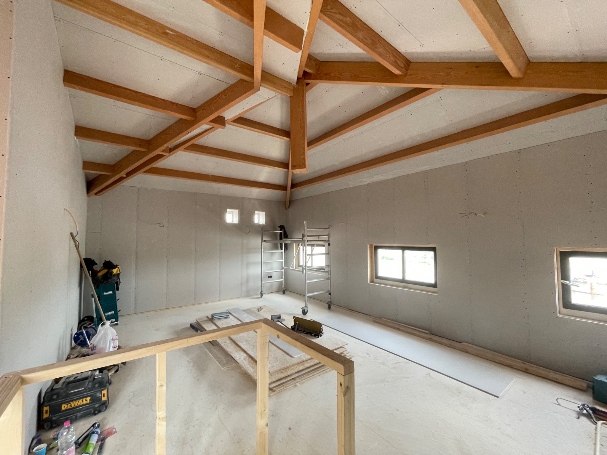 Wnętrze w trakcie wykańczania: surowe ściany z płyt, drewniana konstrukcja dachu, rusztowanie, narzędzia i materiały budowlane porozrzucane na podłodze.