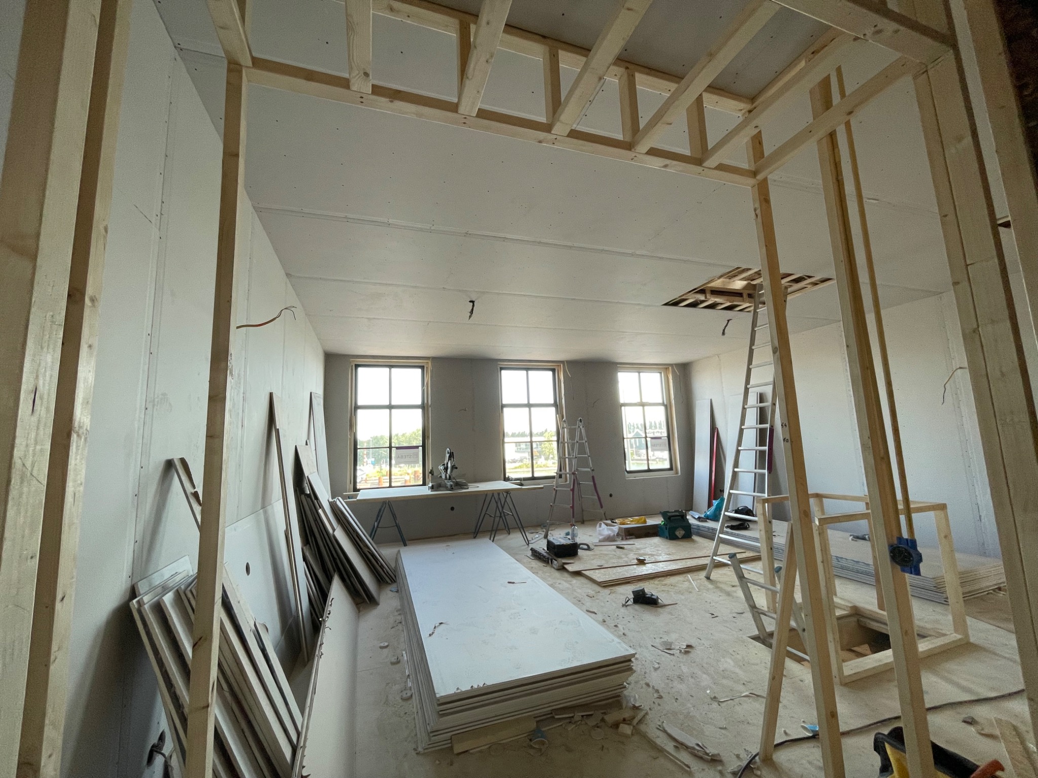 Wnętrze w trakcie remontu: widoczne drewniane stelaże, ułożone płyty na podłodze, ściany częściowo obłożone płytami, okna w tle i drabina. Otwór w suficie z widoczną konstrukcją.