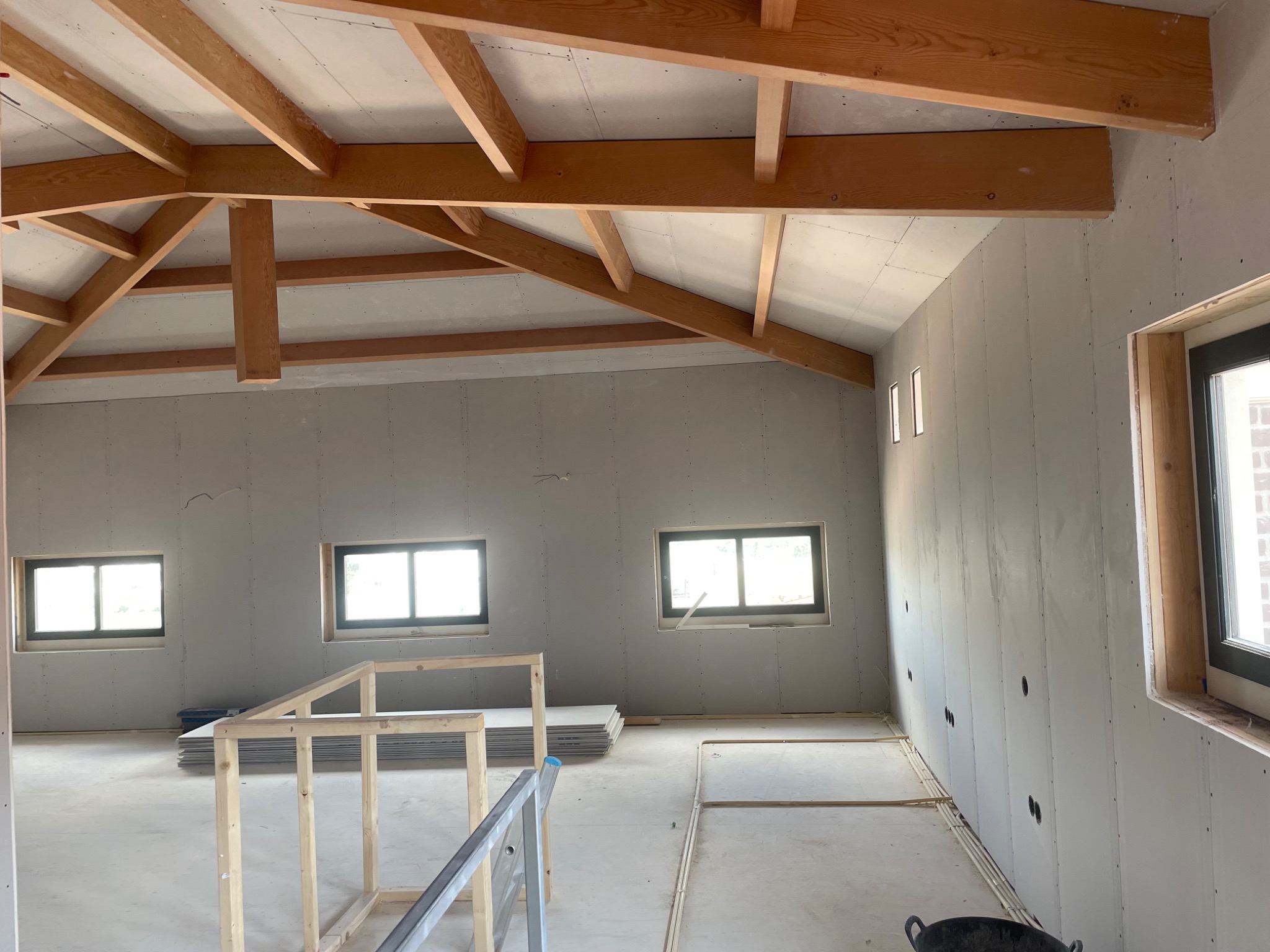 Wnętrze w trakcie montażu okładzin ściennych, widoczne drewniane krokwie stropowe, ułożone przewody elektryczne w podłodze i stos arkuszy materiału budowlanego w rogu pomieszczenia.