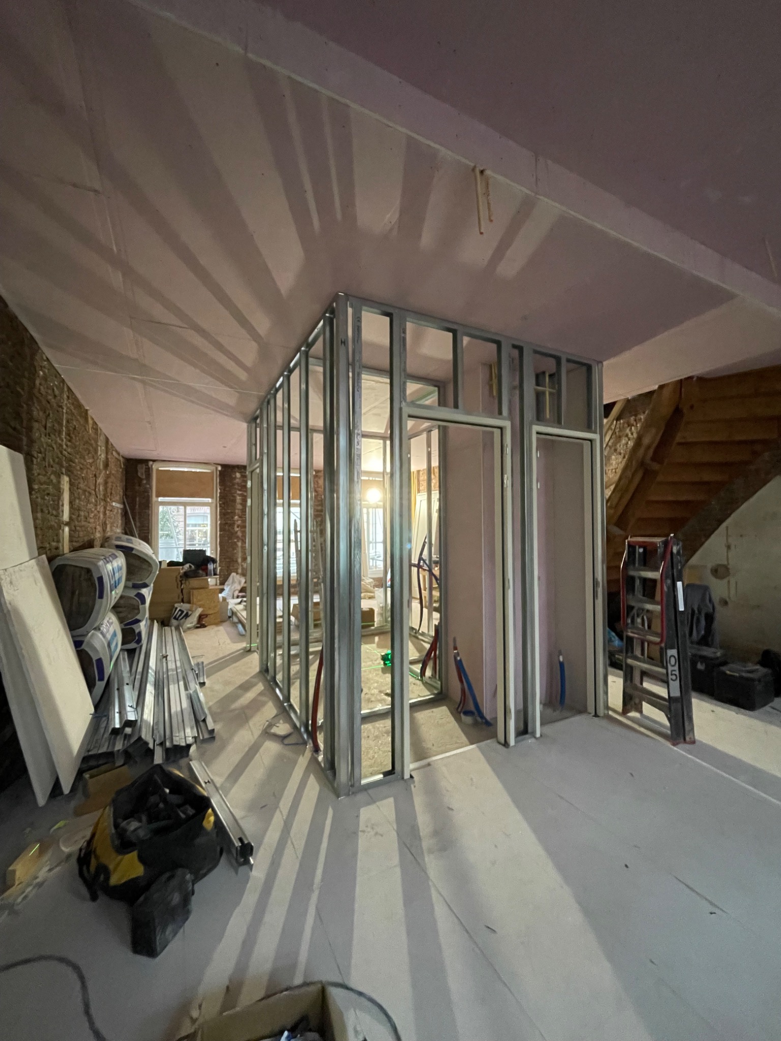 Wnętrze w trakcie remontu: widoczne metalowe profile ścian działowych, przygotowane płyty, narzędzia, sufit i ściany w trakcie wykańczania. Widoczne również schody. Oświetlenie dzienne wpada przez...