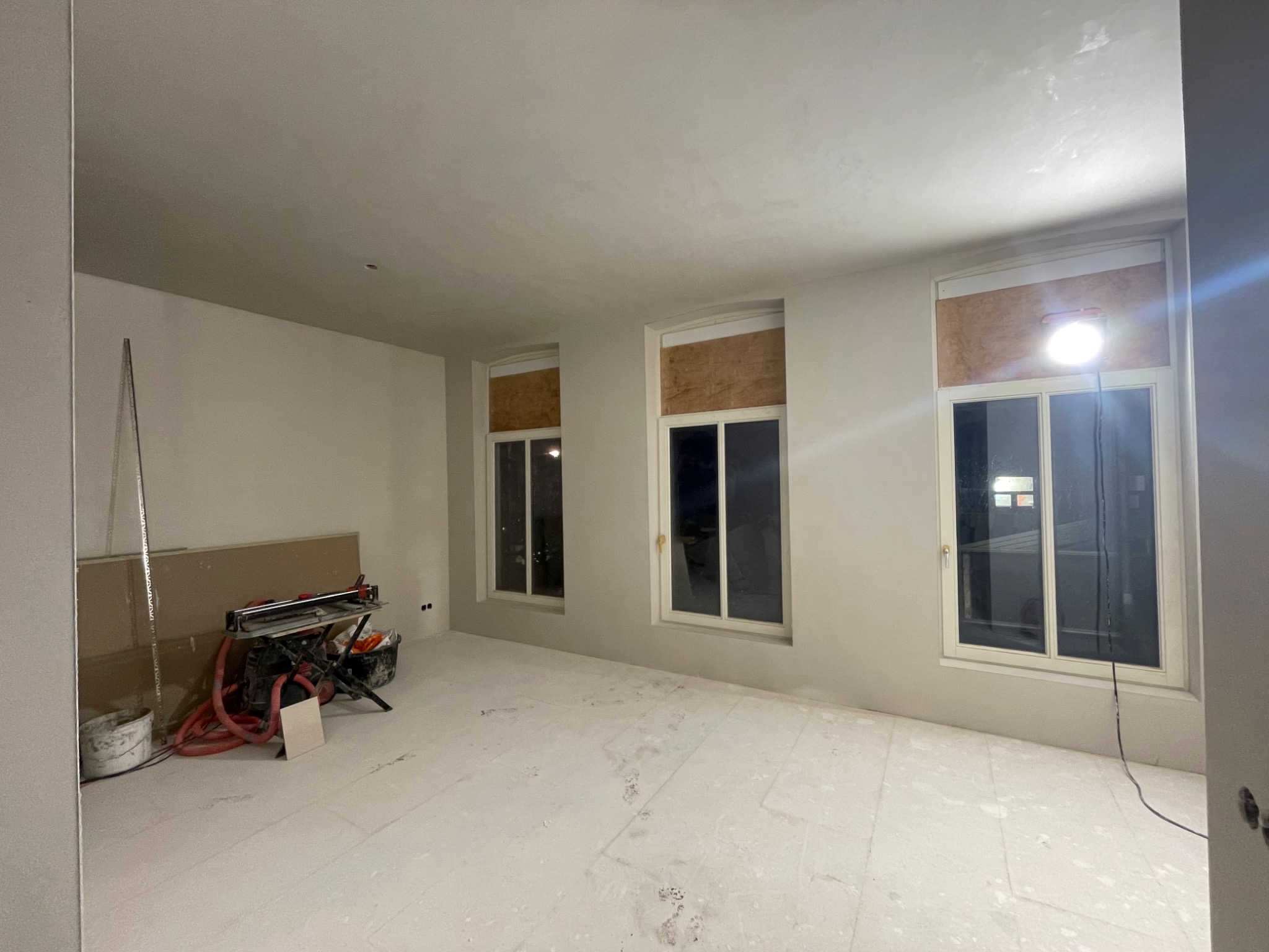 Pokój w trakcie remontu: surowe ściany, zabezpieczone okna, narzędzia do cięcia płyt kartonowo-gipsowych i płyty opierające się o ścianę.