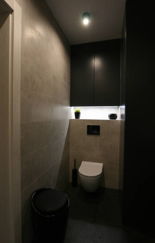 Nowoczesna toaleta z szarymi płytkami, podświetlanymi szafkami i czarnymi akcentami, w tym koszem i szczotką toaletową.