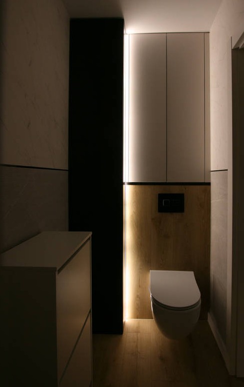 Nowoczesna łazienka z podwieszaną toaletą, szafką pod umywalką i oświetleniem LED wzdłuż ściany, wykończona w odcieniach bieli, czerni i drewna.