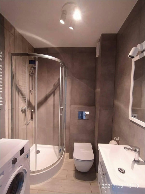 Wykończona łazienka z kabiną prysznicową, podwieszaną toaletą, umywalką i pralką w odcieniach szarości, oświetlona trzema reflektorami punktowymi.