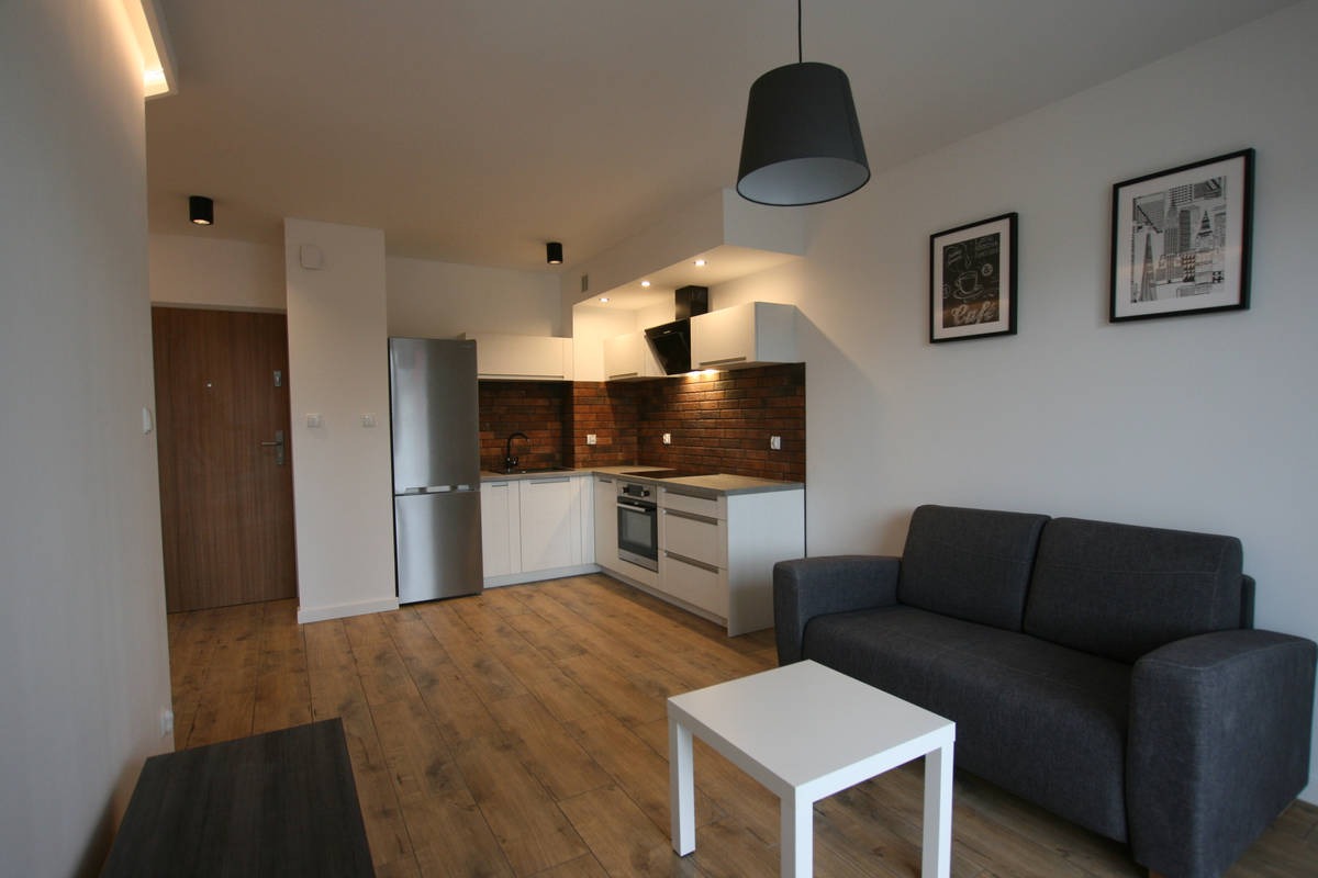 Nowoczesne wnętrze mieszkania z aneksem kuchennym, ceglaną ścianą nad blatem, szarą sofą i białym stolikiem kawowym.