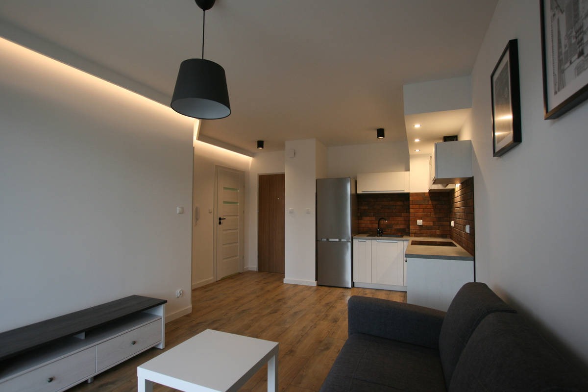 Salon z aneksem kuchennym po remoncie, widoczne podświetlenie LED na suficie, szara sofa i drewnopodobna podłoga.