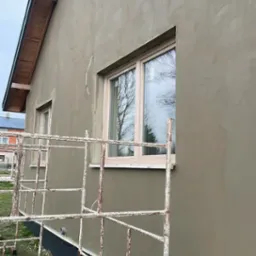 Świeżo otynkowana ściana zewnętrzna domu jednorodzinnego w kolorze beżowym, widoczne rusztowanie, okno z białymi ramami i fragment dachu z drewnianą podbitką.