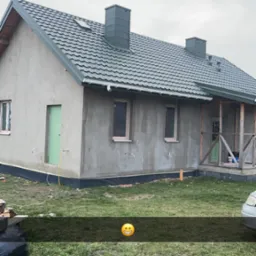 Dom jednorodzinny w trakcie prac elewacyjnych: surowa szara elewacja, zielone drzwi wejściowe, szara dachówka, widoczne przewody i rurki instalacyjne przy ścianach, trawnik przed budynkiem...