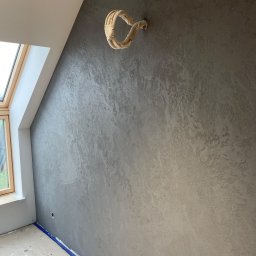 budmaxpainter Jakub Budnik - Ściana w pokoju na poddaszu z dekoracyjną szarą strukturą, zabezpieczone rury grzewcze, okno dachowe i taśma malarska na podłodze.