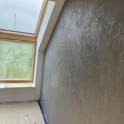 budmaxpainter Jakub Budnik - Ściana z dekoracyjną, szarą fakturą w pokoju z oknem dachowym i zabezpieczoną podłogą, widoczne przewody pod sufitem.