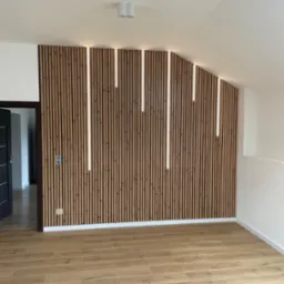 Drewniana ściana z pionowymi listwami i wbudowanym oświetleniem LED, widoczne gniazdka elektryczne, podłoga wyłożona panelami imitującymi drewno, drzwi wejściowe po lewej stronie.