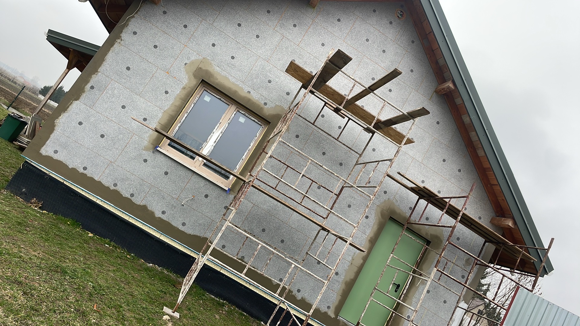 Dom w trakcie ocieplania styropianem, widoczne rusztowanie, okno z drewnianą ramą i zielone drzwi wejściowe, na elewacji szare płyty styropianowe z czarnymi kołkami.